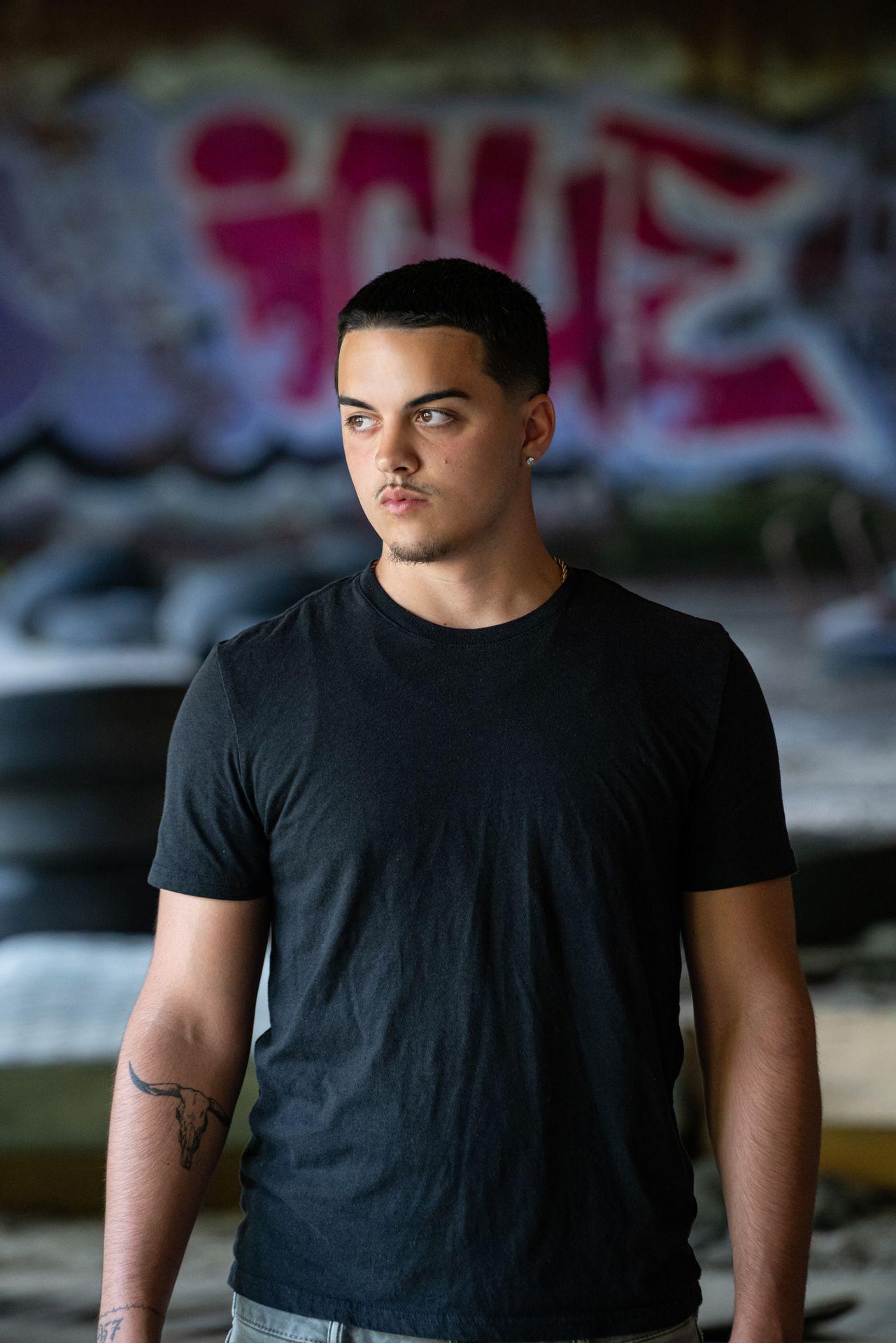 Young man in black t-shirt standing in front of graffiti wall, with tattoo on forearm.