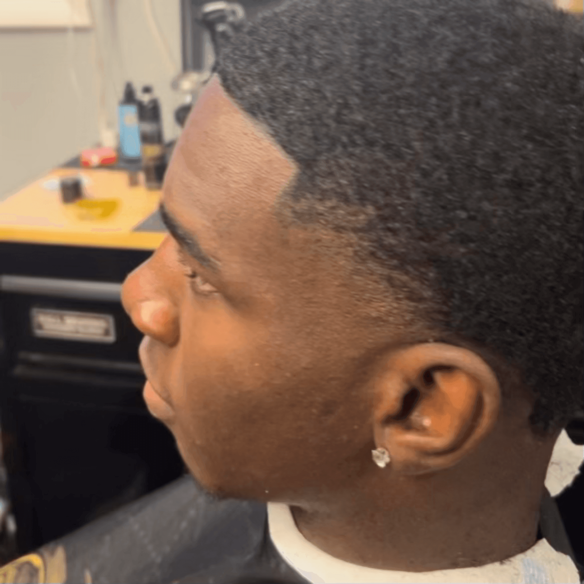 Person with short curly hair sitting in a barber chair, wearing a black and gold salon cape.