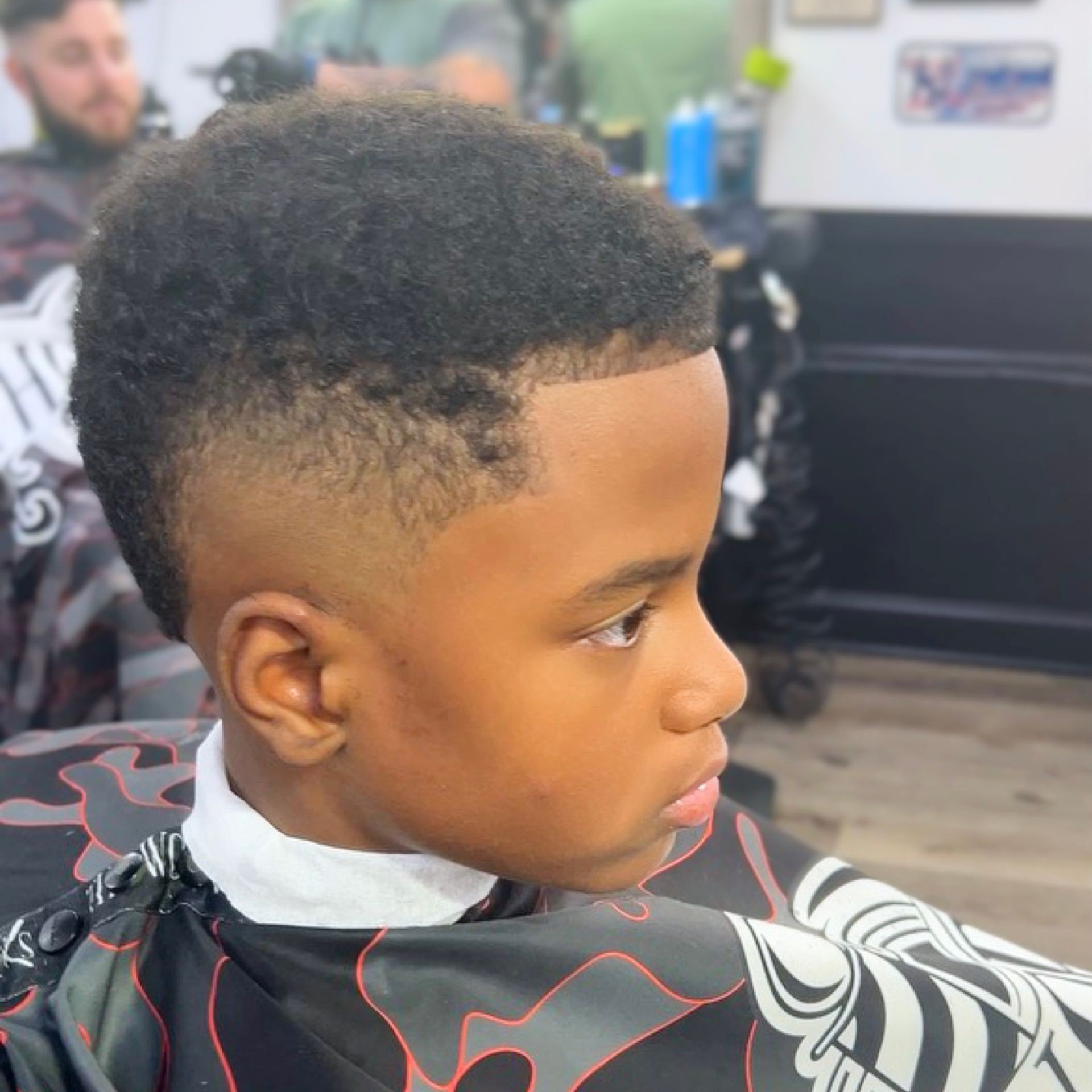 Young boy with a fresh haircut sitting in a barber chair, wearing a patterned cape.